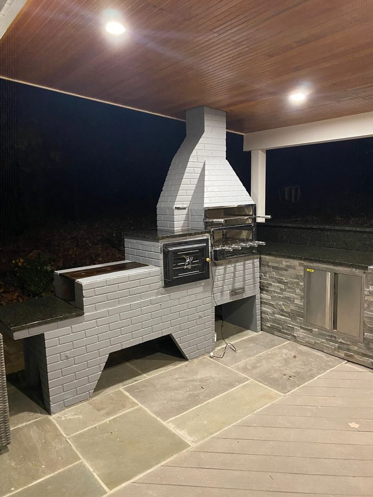 Churrasqueira branca e cinza instalada em área coberta, com forno lateral e fogão.